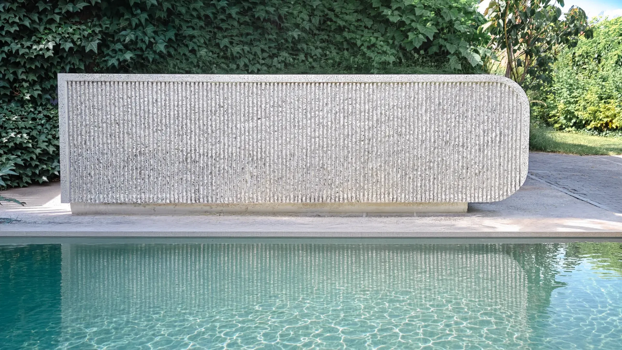 The ribbed front face of ISF 002, a cast concrete volume with a rounded end, seen across the water of a swimming pool
